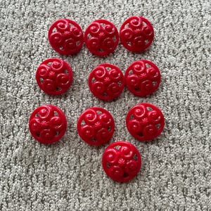 Może przedstawiać: Dziesięć jasnoczerwonych, okrągłych guzików z dekoracyjnym, wirującym wzorem, ułożonych na szarej, teksturowanej powierzchni. Każdy guzik ma średnicę około 2,5 cm. Guziki są równomiernie rozmieszczone.