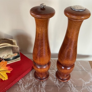 Op de afbeelding: Twee houten zout- en pepermolens met een glanzende afwerking. De zoutmolen heeft een zilveren bovenkant met gaatjes, terwijl de pepermolen een zilveren knop heeft. De molens zijn hoog en hebben een bolvorm.