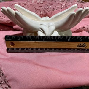 May include: A white ceramic decorative bowl in the shape of two hands holding a central area. The bowl is approximately 6 inches wide. A wooden ruler with inch measurements is placed in front of the bowl.