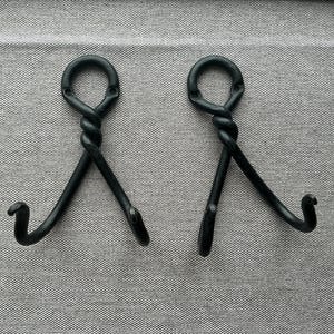 May include: A pair of black, hand-forged metal hooks. Each hook has a twisted loop at the top and a curved hook at the bottom. The hooks are designed for hanging items and are set against a textured grey background.