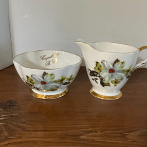 Peut inclure: Un ensemble sucrier et crémier en porcelaine blanche. L'ensemble présente un motif floral avec des fleurs blanches, des feuilles vertes et des centres rouges. Des bordures dorées ornent les bords. Le sucrier porte le mot "Coquille" en lettres dorées.