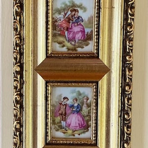 Peut inclure: Un cadre mural rectangulaire avec une finition dorée ornée contenant deux carreaux de porcelaine superposés. Chaque carreau présente une scène peinte colorée de deux personnages en costumes d'époque dans un paysage avec des arbres.