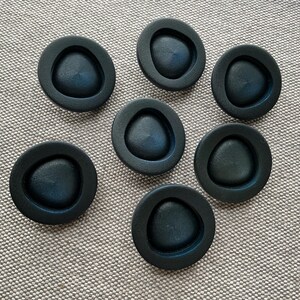 Peut inclure: Sept poignées de meubles noires et circulaires avec un centre triangulaire en retrait. Les poignées sont disposées sur une surface texturée de couleur neutre. La quincaillerie est probablement destinée aux meubles ou aux armoires.