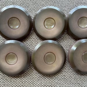 May include: Six brown, circular objects with a central, lighter-colored circle. The objects have a slightly raised edge and a smooth surface. They are arranged in two rows of three on a textured, neutral-colored surface.