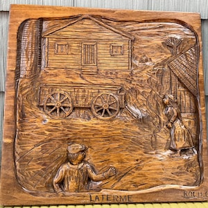 Puede incluir: Una placa de pared cuadrada de madera con un relieve tallado a mano de una escena de granja. La talla muestra una casa, un carro con dos ruedas, una persona de pie y una persona arrodillada. El texto LA FERME y la firma ROCH.C están tallados en la parte inferior.