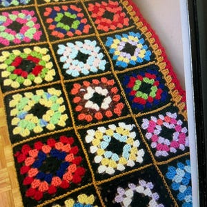 May include: A vibrant, hand-crocheted granny square blanket. The blanket has a patchwork design with squares in various colours, including pink, green, blue, and red, set against a black background. The edges are finished with a red border and gold trim.