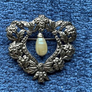 Peut inclure: Broche argentée avec un motif floral en forme de cœur. Une fausse perle en forme de poire pend du centre. La broche présente un nœud en bas et un petit strass en haut.