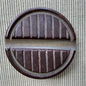 Puede incluir: Un cenicero circular marrón oscuro con un diseño acanalado. El cenicero tiene un área empotrada para apoyar cigarrillos y un borde elevado. El diseño es simétrico, con crestas verticales en las mitades superior e inferior.