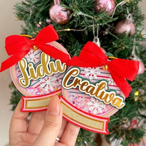 Puede incluir: Dos adornos navideños con lazos rojos se sostienen frente a un pequeño árbol de Navidad. Los adornos son redondos con letras doradas y un fondo a rayas rosas y blancas. Los adornos tienen las palabras "Lidu" y "Creativa".