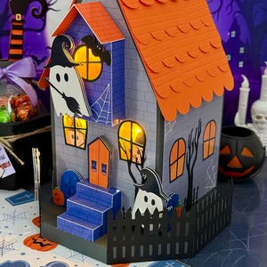 Puede incluir: Una casa de papel con temática de Halloween con techo naranja, paredes azules y recortes decorativos de fantasmas, murciélagos y calabazas. La casa tiene una valla negra y una escalera azul. Incluye el texto "Liducreativa".