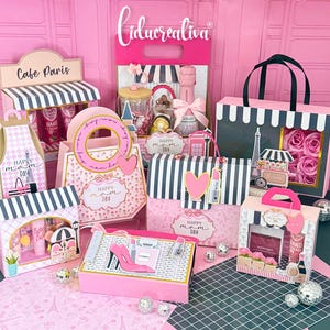 Op de afbeelding: Een verzameling roze en witte geschenkdozen en tassen met een Parijs thema. De dozen hebben strepenpatronen, hartaccenten en de tekst "Happy Mom Day". Sommige dozen hebben Eiffeltoren- en café-ontwerpen.