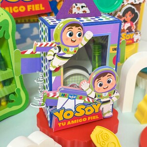 May include: A rectangular purple and blue paper box featuring cartoon space ranger characters, green accents, and the text YO SOY TU AMIGO FIEL. The box has a handle on top and a clear window, sitting on a red scalloped base with yellow plastic shapes.