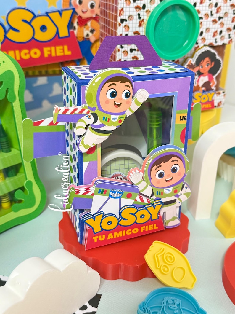 May include: A rectangular purple and blue paper box featuring cartoon space ranger characters, green accents, and the text YO SOY TU AMIGO FIEL. The box has a handle on top and a clear window, sitting on a red scalloped base with yellow plastic shapes.