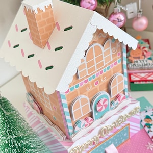 Puede incluir: Una casa de jengibre de papel con techo blanco, chimenea y adornos decorativos. La casa presenta ventanas arqueadas, detalles de caramelos y una base con un patrón de rayas rosas y blancas. Las palabras "Merry Christmas" son visibles.