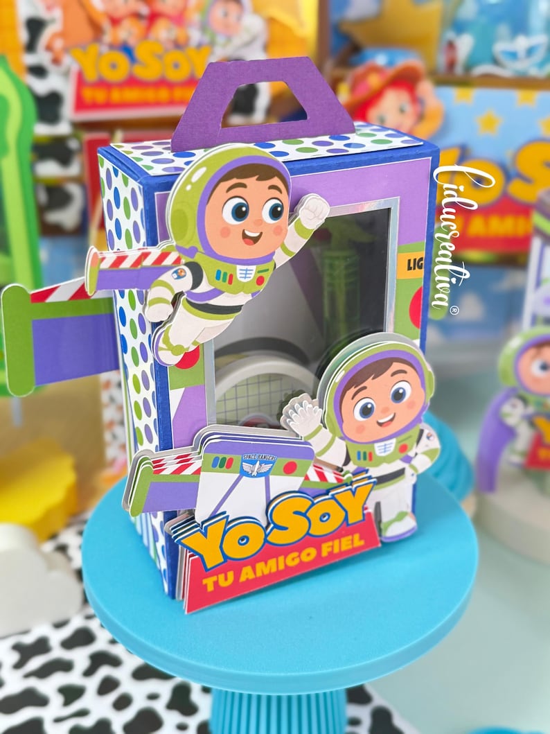 May include: A rectangular purple and blue cardboard favour box featuring a cutout handle and a clear window. The box is decorated with layered paper cutouts of a cartoon space ranger character and the text Yo Soy Tu Amigo Fiel in yellow and red.