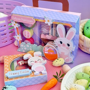 Puede incluir: Cajas de regalo con temática de Pascua con decoraciones de conejos y huevos. Una caja tiene la inscripción "Felices Pascuas". Otra caja tiene la inscripción "Happy Easter". También hay una zanahoria y huevos moteados.