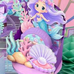 Può includere: Un centrotavola in carta ritagliata a più strati raffigurante una sirena dei cartoni animati con capelli viola e una corona di fiori, circondata da corallo rosa, rocce color sabbia e una conchiglia viola con una perla bianca su base.