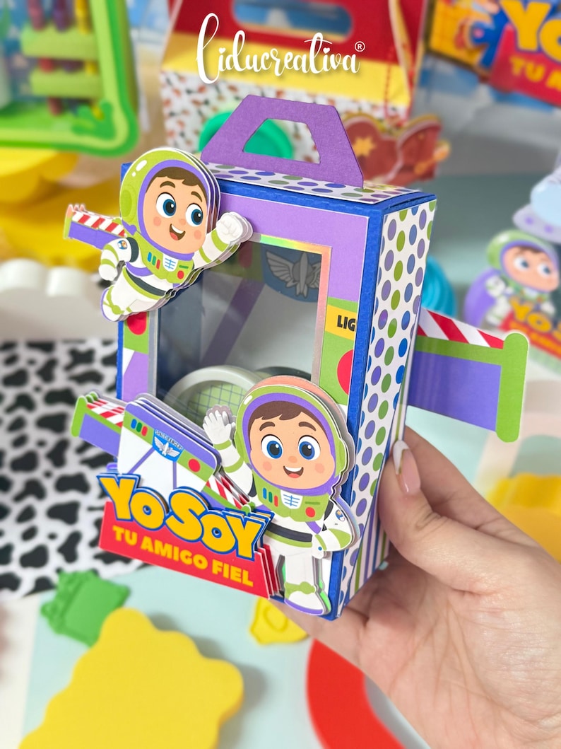 May include: A rectangular purple and blue cardboard box with a handle, featuring cartoon space ranger characters in white, green, and purple suits. The box has a clear window and displays the text Yo Soy Tu Amigo Fiel in yellow and blue letters.
