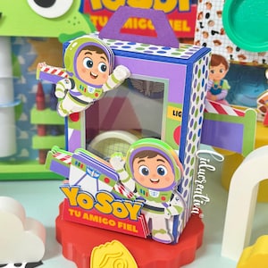 Pode incluir: Uma caixa de presente retangular de papel com recortes do personagem Buzz Lightyear, uma alça roxa e painéis laterais com bolinhas azuis e brancas. A frente exibe o texto Yo Soy Tu Amigo Fiel com uma janela transparente.