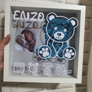 Può includere: Una scatola ombra con cornice bianca con il nome "ENZO" in lettere bianche. All'interno, una foto di un bambino, un orso pieno di rose blu e dettagli della nascita: 3.350 kg, 50 cm e la data e l'ora di nascita.