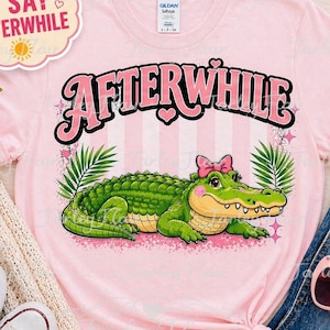 May include: A light pink t-shirt with the text "AFTERWHILE" in pink and black, featuring a cartoon alligator with a pink bow. The alligator is green with a yellow belly, surrounded by green leaves and pink stars.