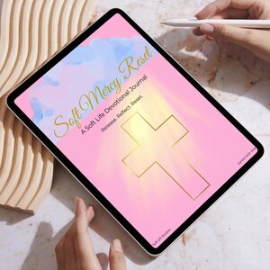 Può includere: Un tablet digitale che mostra un diario devozionale intitolato "Soft Mercy Reset". Lo schermo presenta uno sfondo rosa con una croce dorata e testo. Il sottotitolo del diario è "A Soft Life Devotional Journal". Uno stilo è tenuto vicino allo schermo.