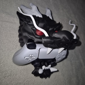 Könnte beinhalten: Eine schwarz-weiße Drachenfigur, die einen grauen Videospiel-Controller hält. Der Drache hat rote Augen und weiße Akzente. Der Controller hat eine graue Oberfläche mit silbernen Knöpfen.