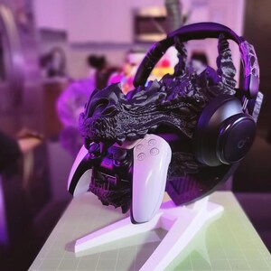 Könnte beinhalten: Ein schwarzer Kopfhörerständer in Drachenform, der einen schwarzen Kopfhörer und einen weißen Gamecontroller hält. Der Ständer steht auf einem weißen Sockel. Der Drachenkopf ist mit strukturierten Schuppen und scharfen Zähnen versehen.