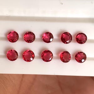 Könnte beinhalten: Zehn runde, facettierte, tiefrote Edelsteine sind in zwei Reihen auf einer weißen Oberfläche angeordnet. Die Edelsteine sind gleichmäßig verteilt und scheinen von ähnlicher Größe und Klarheit zu sein. Die Edelsteine haben eine lebendige, gesättigte Farbe.