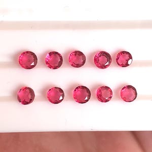 Może przedstawiać: Dziesięć okrągłych, fasetowanych, jasno czerwonych kamieni szlachetnych ułożonych w dwóch rzędach na białej powierzchni. Kamienie mają wysoki połysk i wydają się być podobnej wielkości i kształtu.