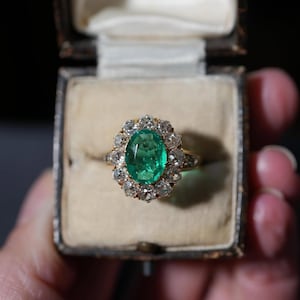 Könnte beinhalten: Ein goldener Ring mit einem ovalen Smaragd, umgeben von einem Diamanten-Halo. Der Ring wird in einer Vintage-Schmuckschatulle mit cremefarbenem Innenraum präsentiert. Der Smaragd ist ein leuchtendes Grün, die Diamanten funkeln.