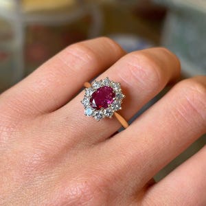 Może przedstawiać: Złoty pierścionek z owalnym rubinem otoczonym przez otoczkę z diamentów i akwamarynów. Złota obrączka podkreśla głęboką czerwień rubinu i blask otaczających kamieni. Piękna biżuteria.