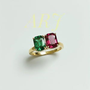 Könnte beinhalten: Ein Goldring mit zwei nebeneinander liegenden Edelsteinen im Smaragdschliff, einer grün und einer pink, wobei die Buchstaben ARJ in hellgelbem Text über dem Ring stehen. Die Ringschiene besteht aus poliertem Gold mit schlichtem Design.