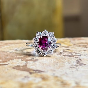 Könnte beinhalten: Ein silberner Ring mit einem großen, oval geschliffenen, tiefrosa Edelstein, umgeben von einem Halo aus kleineren, klaren Edelsteinen. Der Ring ist vor einem neutralen Hintergrund platziert, der das Funkeln der Steine hervorhebt.