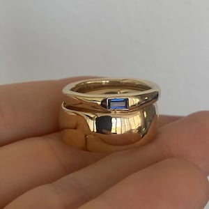 Puede incluir: Dos anillos de oro, uno apilado sobre el otro. El anillo superior tiene una gema azul rectangular. Los anillos son lisos y brillantes, reflejando la luz. Los anillos están sobre un fondo neutro.