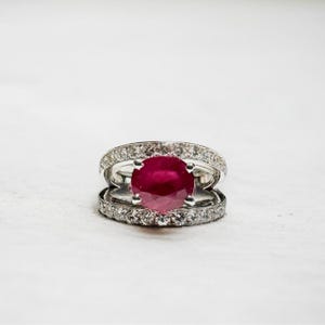 Peut inclure: Une bague en argent ornée d'un grand rubis rouge foncé de forme ovale. Le rubis est entouré d'un halo de petits diamants transparents. La monture est également sertie de petits diamants. L'ensemble est présenté sur fond blanc.