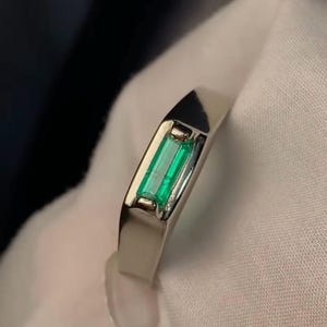 Puede incluir: Un anillo de plata con una gema verde rectangular con talla esmeralda. El anillo tiene un diseño elegante y moderno con un acabado pulido. La gema está engastada en un bisel, realzando su color y forma vibrantes. El anillo se muestra sobre una superficie blanca.