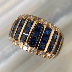 以下が含まれることがあります： 正方形の濃い青色のサファイアの帯と、垂直の金のバーが交互に配置されたゴールドリング。リングは小さく丸いダイヤモンドで装飾されています。このジュエリーはステートメントリングです。