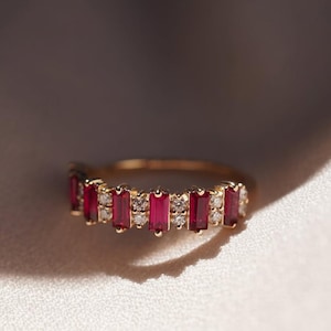 Könnte beinhalten: Ein goldener Ring mit abwechselnd rechteckigen roten Edelsteinen und kleinen, runden, klaren Edelsteinen. Der Ring ist vor einem weichen, weißen Hintergrund mit Schatten platziert.