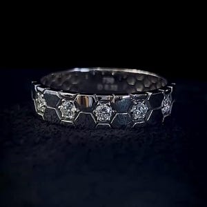 Könnte beinhalten: Ein silberner Ring mit einem Wabenmuster. Der Ring zeigt eine Reihe sechseckiger Formen, von denen jede einen funkelnden Diamanten enthält. Das Band hat eine strukturierte Oberfläche, und der Ring ist vor einem dunklen Hintergrund platziert.