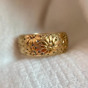 Könnte beinhalten: Ein goldfarbener Ring mit floralem Muster. Der Ring hat ein Band, das mit detaillierten, erhabenen Blumenmustern bedeckt ist. Der Ring ist vor einem weichen, weißen Hintergrund platziert, der die komplizierten Details des Schmucks hervorhebt.