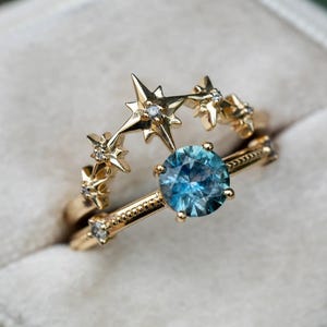 Könnte beinhalten: Zwei goldene Ringe. Ein Ring hat einen großen, runden, hellblauen Edelstein. Der andere Ring hat ein Sterndesign mit kleinen Diamanten. Die Ringe sind vor einem weichen, neutralen Hintergrund platziert.
