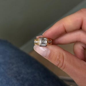 May include: A gold ring with a large, square, clear gemstone as the centerpiece. The ring has a gold band with smaller accent stones on either side of the main stone. The ring is held up by a person's fingers.