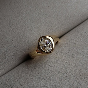 Könnte beinhalten: Ein goldener Ring mit einem funkelnden, oval geschliffenen Diamanten. Der Diamant ist in einer Zargenfassung gefasst, und das Band ist aus glattem, poliertem Gold. Der Ring wird auf einer grauen Oberfläche präsentiert.
