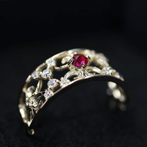 Könnte beinhalten: Ein goldfarbener Ring mit einem verzierten Design, mit einem zentralen roten Edelstein und mehreren kleineren, klaren Steinen. Der Ring hat ein zartes, offenes Muster und ist vor einem dunklen Hintergrund platziert.