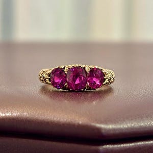 May include: A gold ring featuring three oval, vibrant pink gemstones. The ring has an ornate, filigree design on the band. The gemstones are set in a row, creating a classic and elegant look. The ring is displayed on a brown surface.