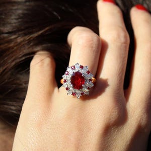 Könnte beinhalten: Ein goldener Ring mit einem großen, ovalen, tiefroten Edelstein, umgeben von kleineren weißen und roten Steinen. Das Design des Rings ist floral, mit dem roten Edelstein im Zentrum. Der Ring befindet sich an einem Finger.