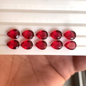 Könnte beinhalten: Zehn facettierte, tropfenförmige, tiefrote Edelsteine sind in zwei Reihen auf einer weißen Oberfläche angeordnet. Die Edelsteine haben einen Brillantschliff, der das Licht reflektiert. Die Edelsteine haben eine leuchtende, gesättigte rote Farbe.