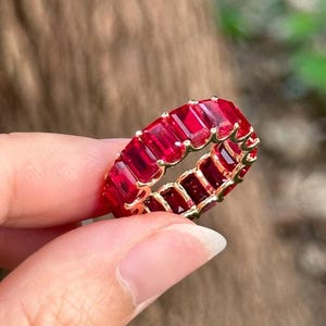 Könnte beinhalten: Ein goldfarbener Ring mit einem durchgehenden Band aus rechteckigen, tiefroten Edelsteinen. Die Edelsteine sind eng aneinander gesetzt und erzeugen eine lebendige Darstellung. Der Ring wird von den Fingern einer Person gehalten, um das Design und die Farbe hervorzuheben.