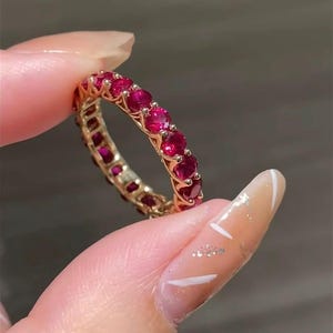 Könnte beinhalten: Ein goldener Ring mit einer durchgehenden Reihe leuchtend roter Edelsteine. Der Ring wird hochgehalten, wodurch das detaillierte Design und die leuchtende Farbe der Steine hervorgehoben werden. Der Ring scheint ein vollständiger Eternity-Ring zu sein.